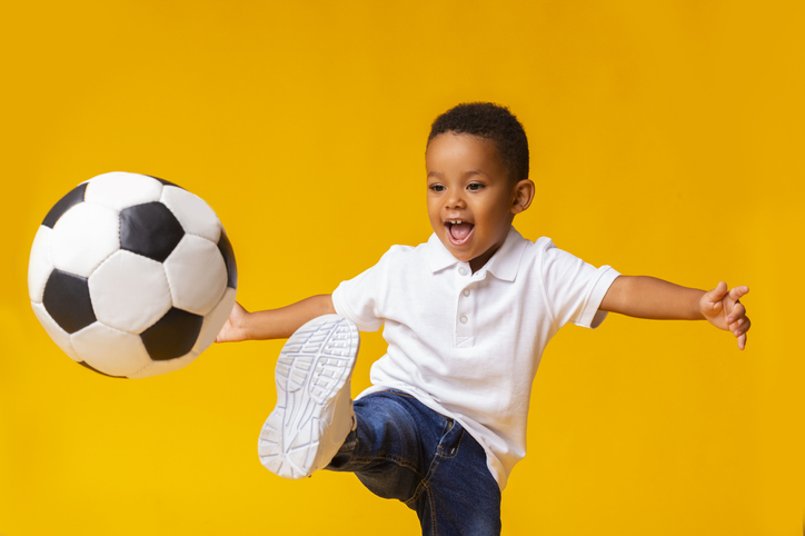 Adorable little boy playing football, hitting ball over yellow background