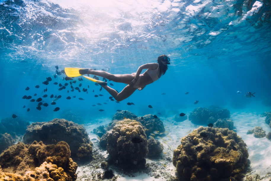 Free diver girl glides with school of fishes in blue ocean
